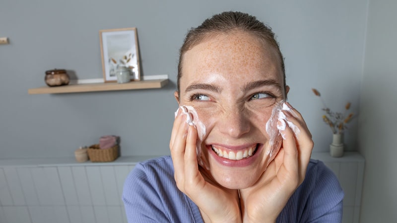 Junge Frau grinst in die Kamera und trägt Creme auf ihr Gesicht auf.