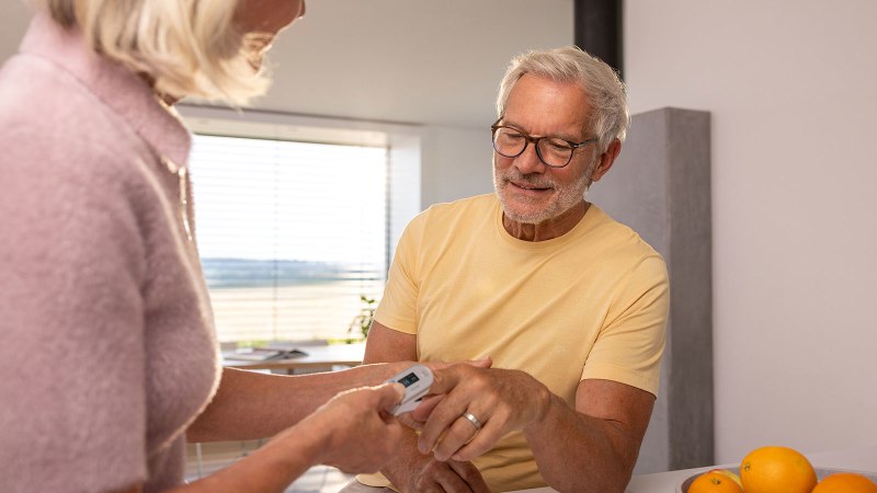 An elderly woman is measuring an elderly man’s oxygen saturation at his home using a Beurer pulse oximeter
