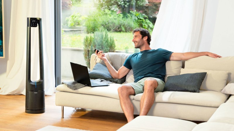 A man is sitting comfortably on the sofa, controlling the LV 500 2-in-1 air purifier and fan with a remote control