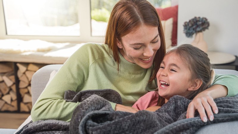Mutter und Tochter sitzen lachend unter einer Wärmezudecke von Beurer auf dem Sofa