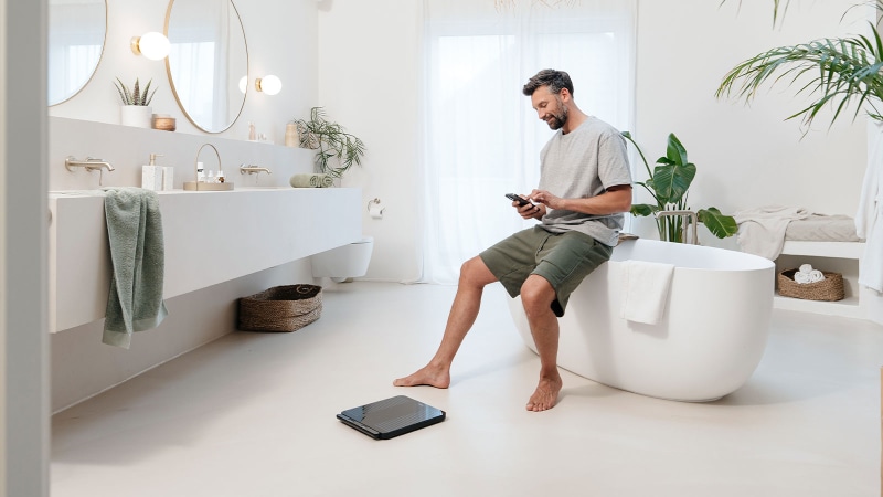 A man is sitting on the edge of the bath and controlling a Beurer diagnostic scale via his smartphone