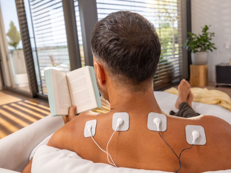 Mann sitzt auf der Couch mit TENS-Elektroden im Nacken und liest ein Buch