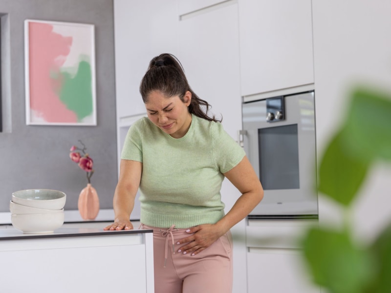 Frau hält sich mit schmerzhaftem Gesichtsausdruck den Unterbauch in einer Küche.