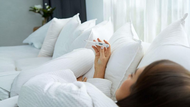 Frau liegt auf der Couch mit einem Fieberthermometer in der Hand