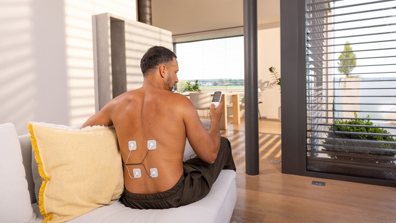 Mann sitzt auf der Couch und hat Elektroden von TENS-Gerät auf dem Rücken 