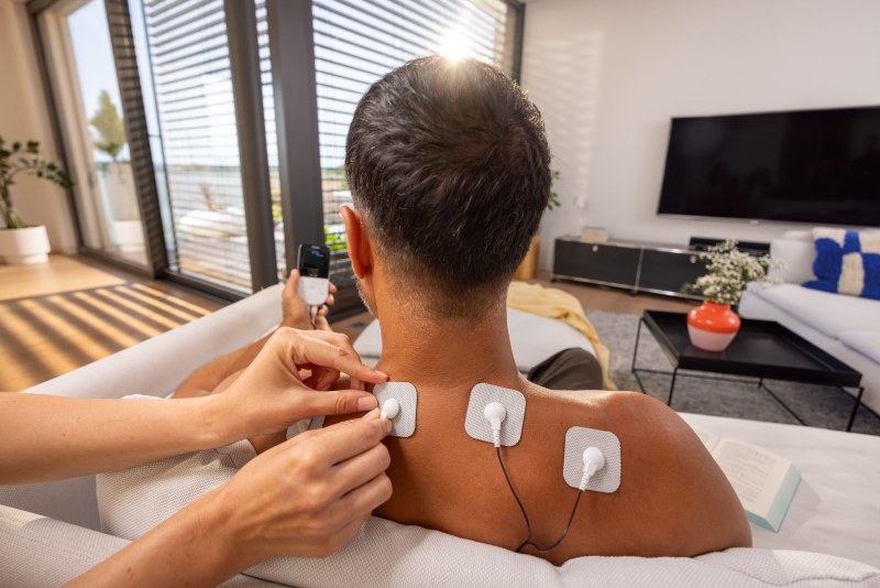 Eine Frau appliziert im heimischen Wohnzimmer ein Elektrodepad des Beurer EM 56 (Digital TENS/EMS) auf den Nackenbereich eines Mannes.