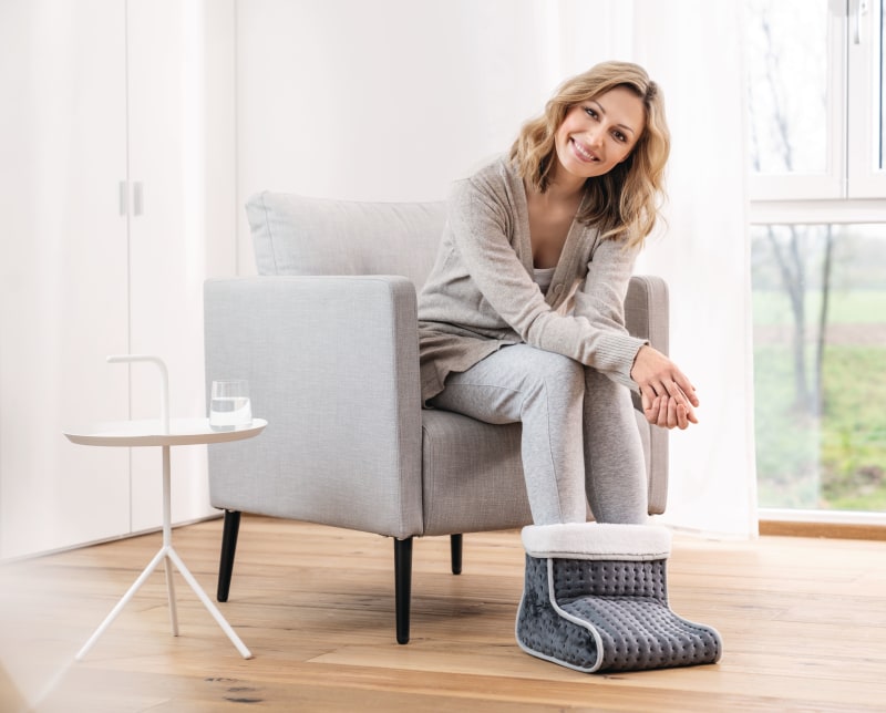 Eine Frau sitzt auf einem weißen Sofa und hat ihre Füße in einem elektrischen Fußwärmer in einem Wohnzimmer.