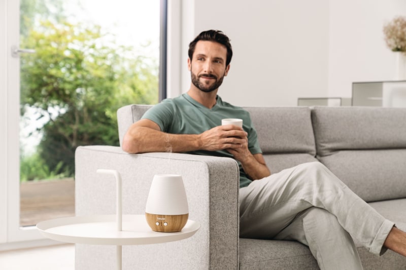 Mann sitzt auf dem Sofa und hat eine Kaffeetasse in der Hand. Im Vordergrund steht ein LA 40 Aroma Diffusor auf einem Beistelltisch.