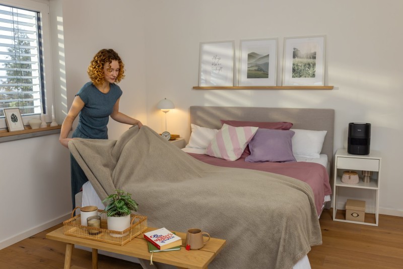 Eine Frau deckt ein Bett mit einer Decke zu. Auf dem Nachttisch steht der Beurer Luftbefeuchter.