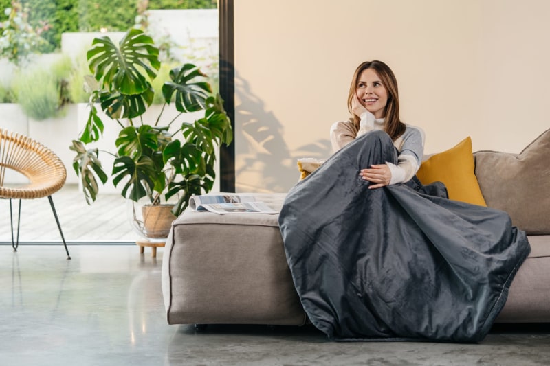 Eine Frau sitzt auf einem Sofa und ist mit einer Heizdecke Beurer zugedeckt. Im Hintergrund sind eine Pflanze und ein Fenster zu sehen.