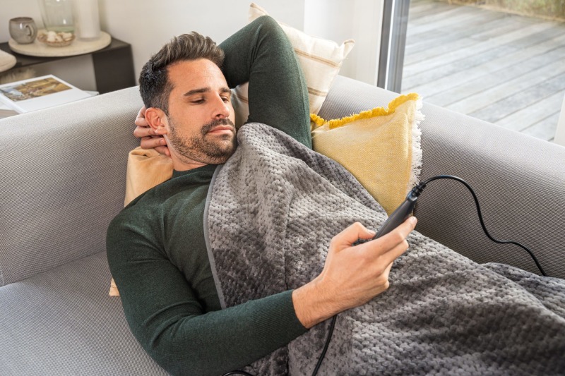 Ein Mann liegt auf einem Sofa und ist mit der Beurer HD 65 Wärmezudecke zugedeckt. Er hält die Fernbedienung der Decke in der Hand.