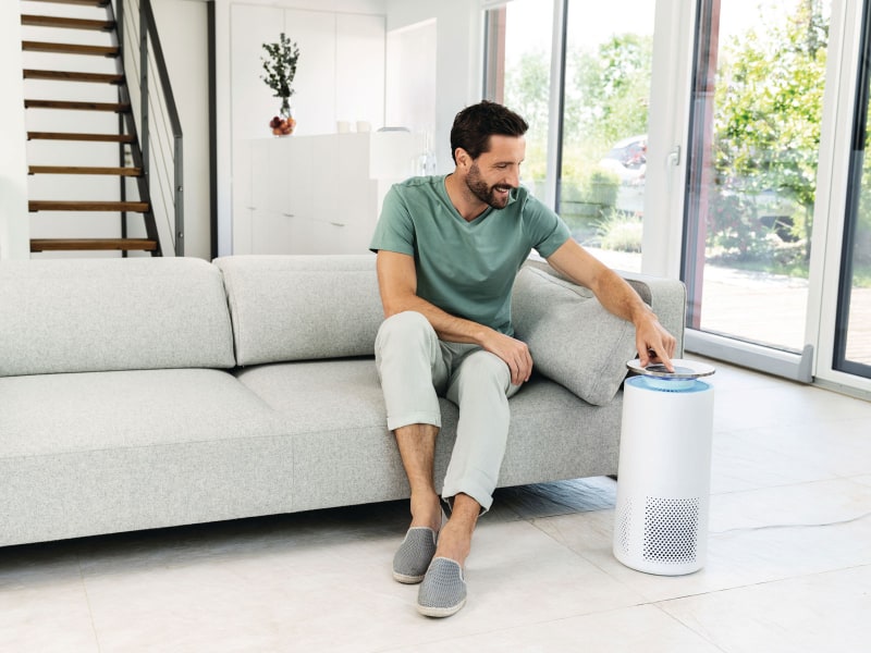 A man sits on a sofa in a living room and operates the Beurer LR 400 air purifier standing next to him.