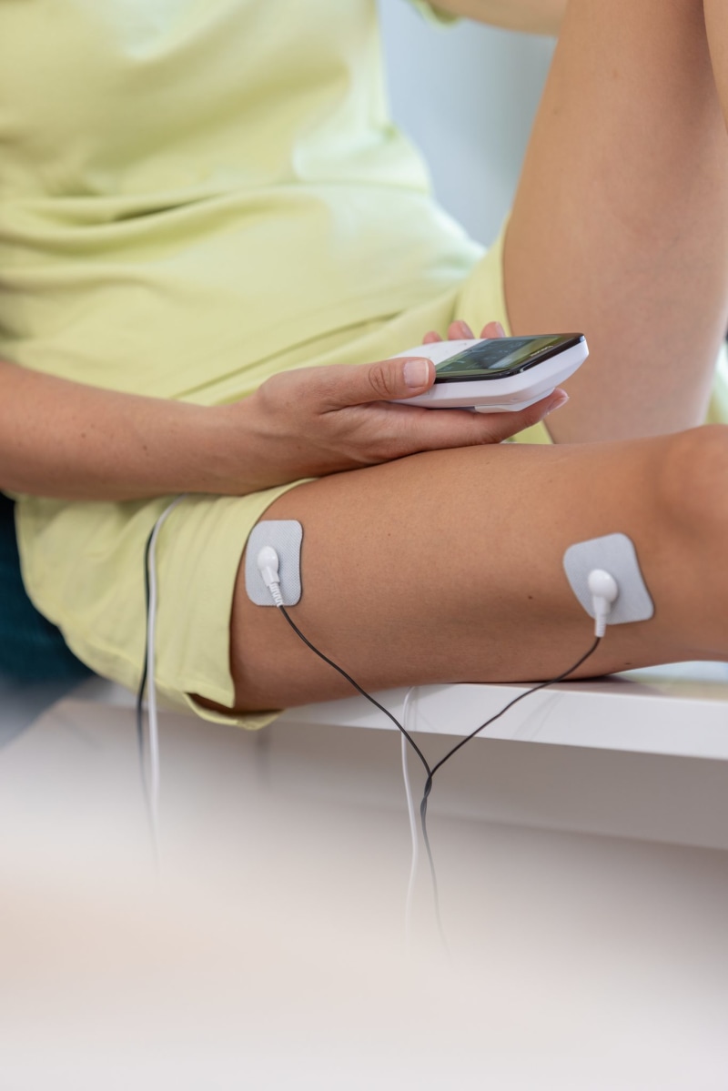 Eine Person hält das Beurer EM 56 (Digital TENS/EMS) in der Hand, während zwei Elektrodenpads mit Kabeln am Oberschenkel befestigt sind.