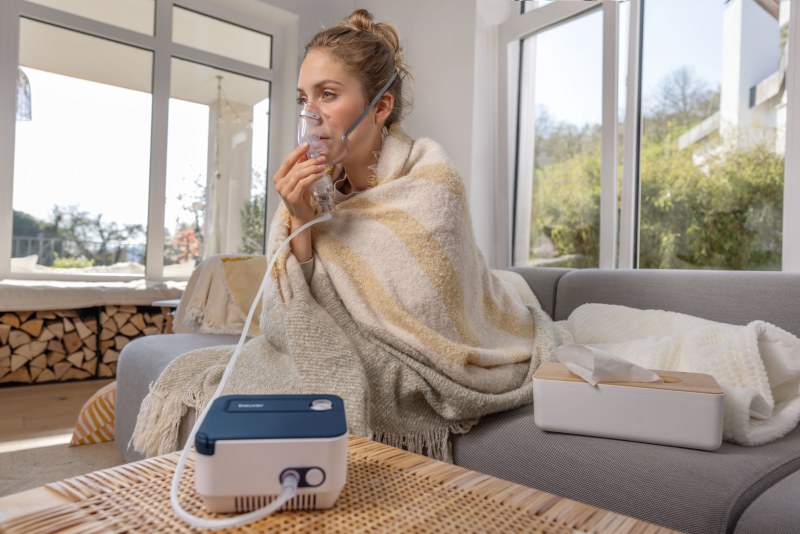 Eine Frau sitzt auf einem Sofa, mit einer Decke zugedeckt, und benutzt den Beurer Inhalator, der auf einem Tisch vor ihr steht. Eine Taschentuchbox liegt neben ihr auf dem Sofa.