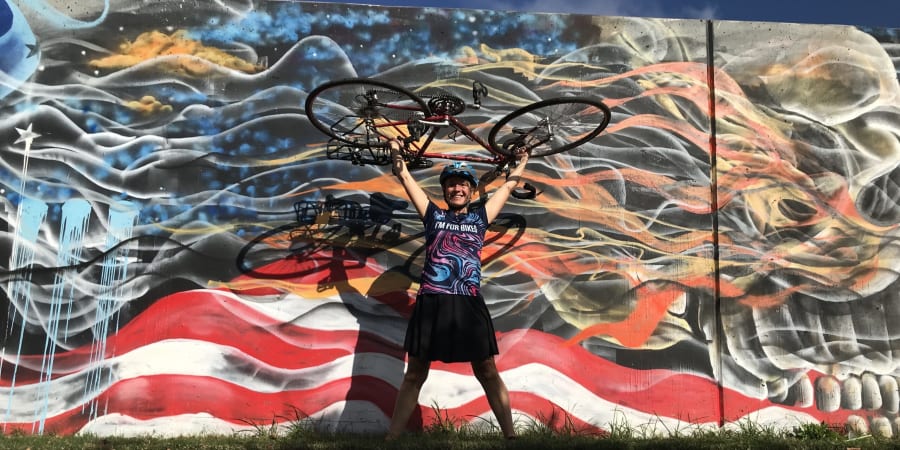 A photograph of Pattie Baker holding her bicycle in front of a mural. 