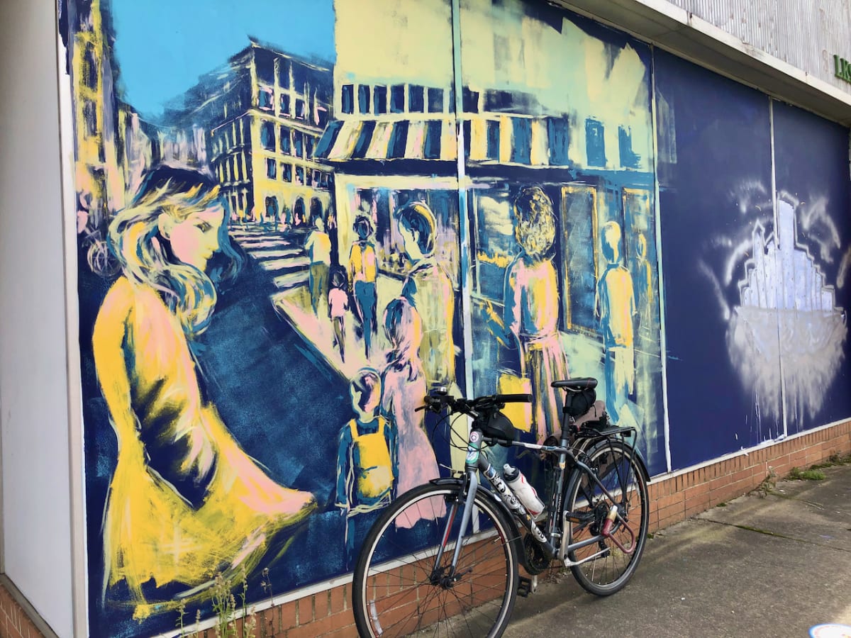 A photo of my bicycle in front of a mural with a painting of a public scene.