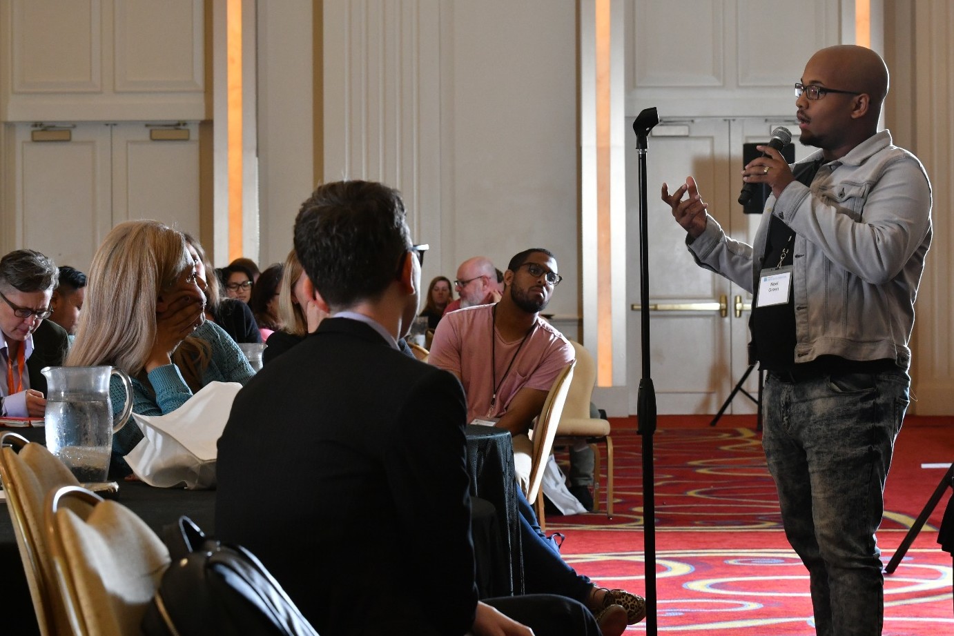 Black masculine presenting person with a blue button up and jeans, speaking into a microphone in front of an audience. Audience is seated around round tables in a ballroom with a red carpet with a yellow pattern.