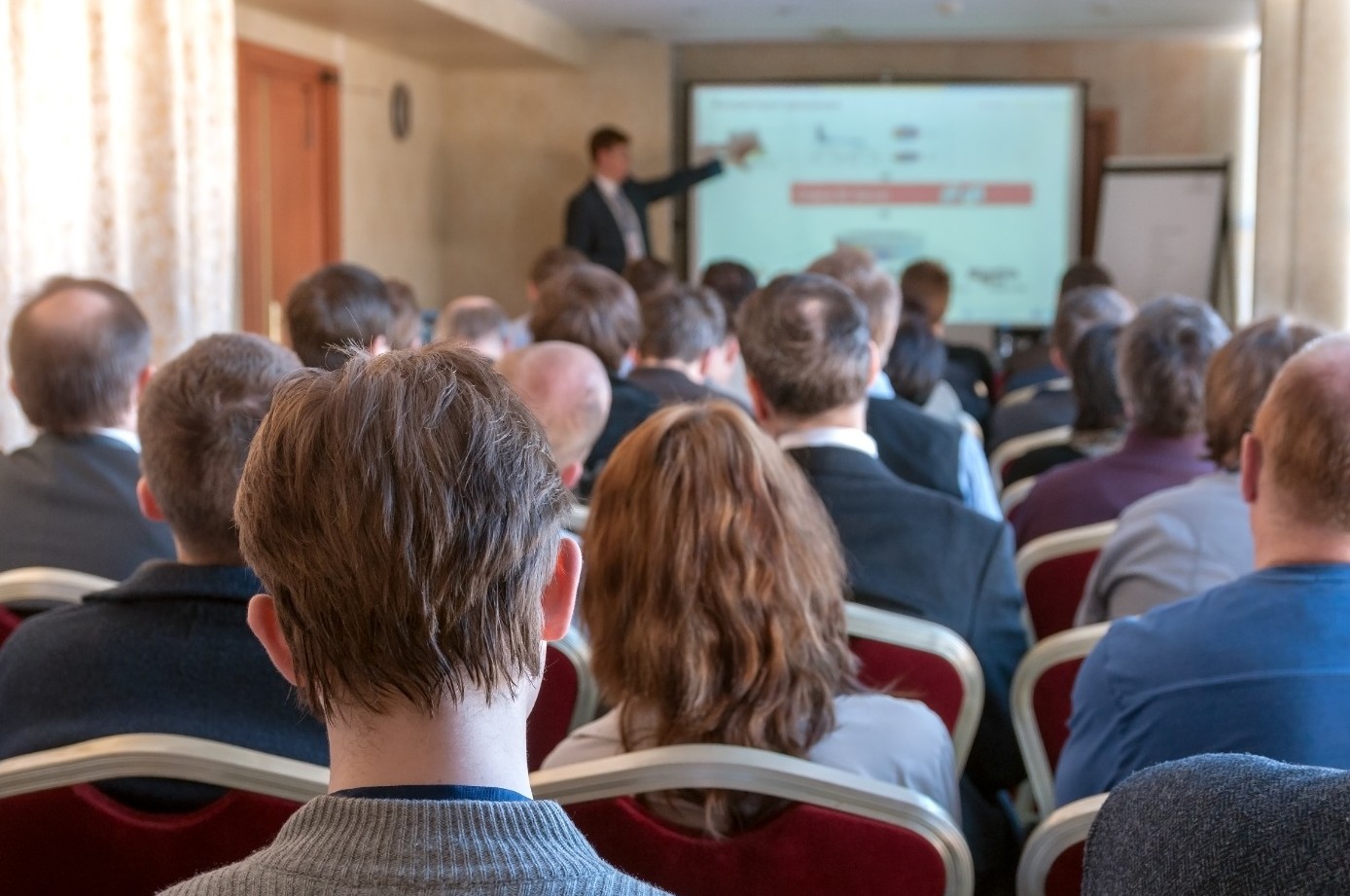 Back view of an audience of people facing a speaker pointing to a presentation on a screen at the front of the room.