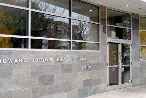 Close up, side view of the front of a building, with large horizontal windows on the left, doors, on the right, and a facade of flagstone and the words "Howard Brown Health Center" and "LCCP"