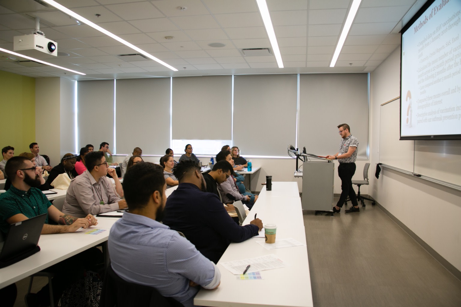 Group of people at long white desks, watching a presenter at a podium in front of a screen at the front of the room.