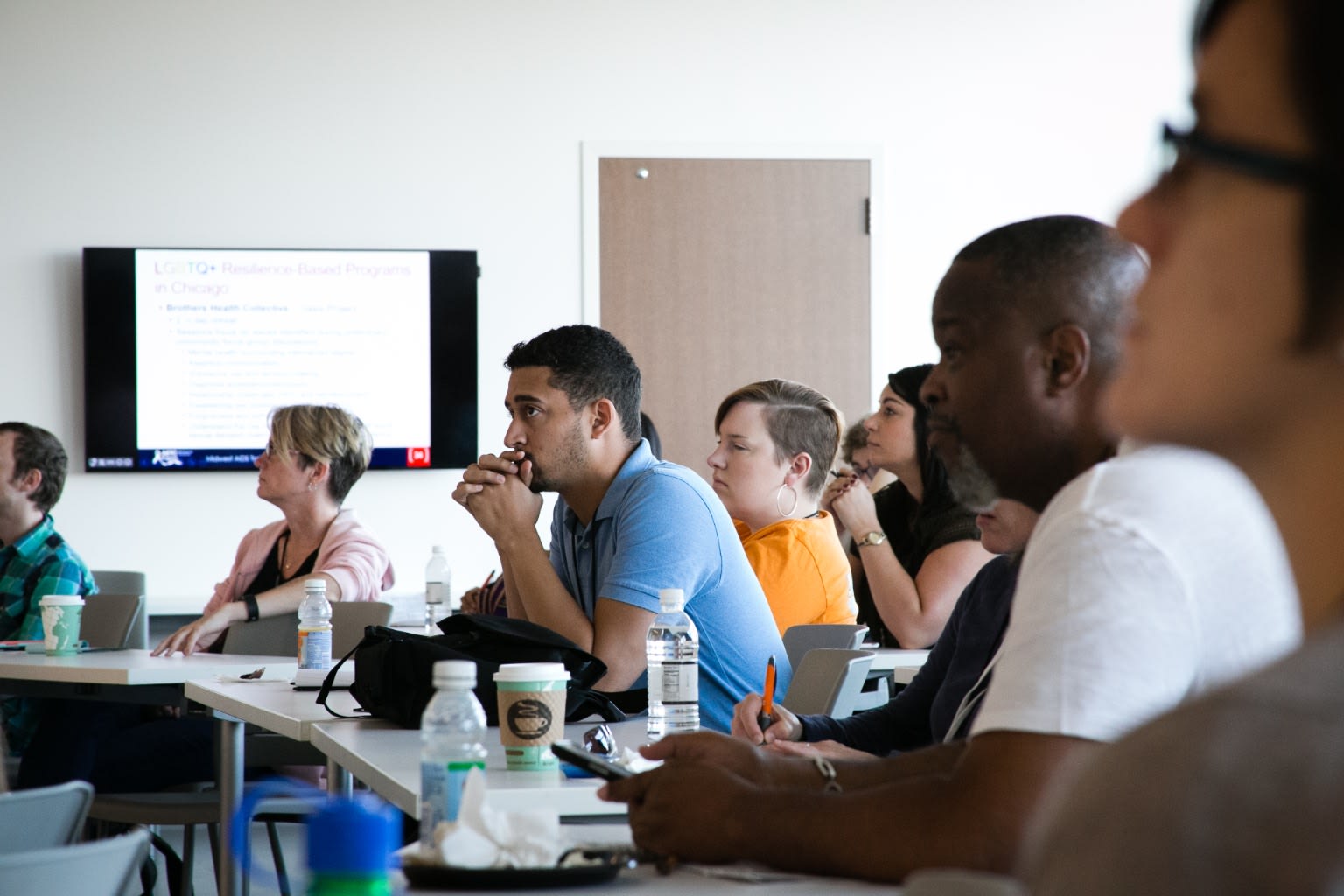 Side view of an audience of about 10 people at long white tables in a classroom, looking towards the front of the room. The desk has water bottles, coffee cups, and some pens.