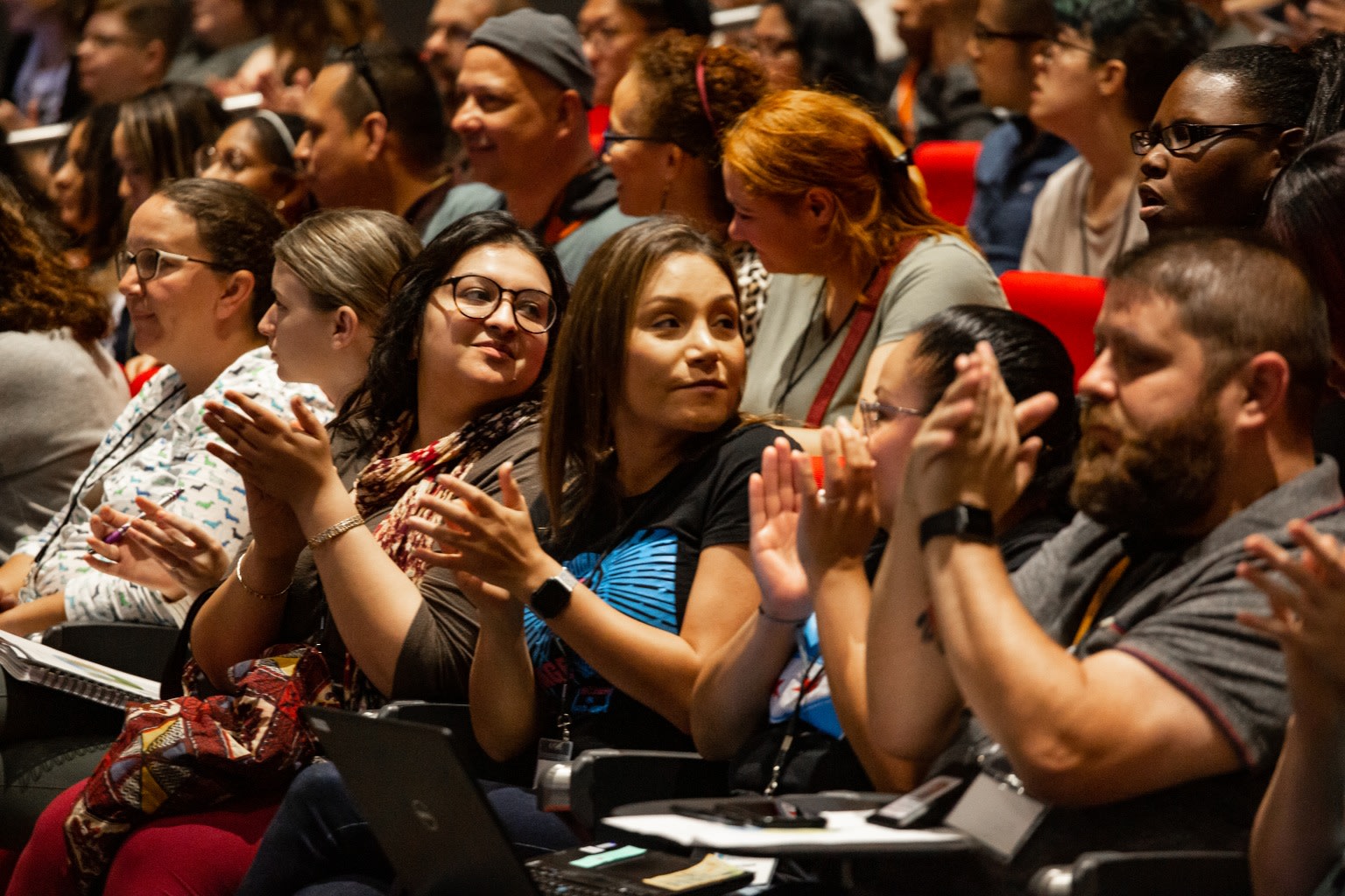 Side view of audience sitting in red chairs in an auditorium and applauding. 