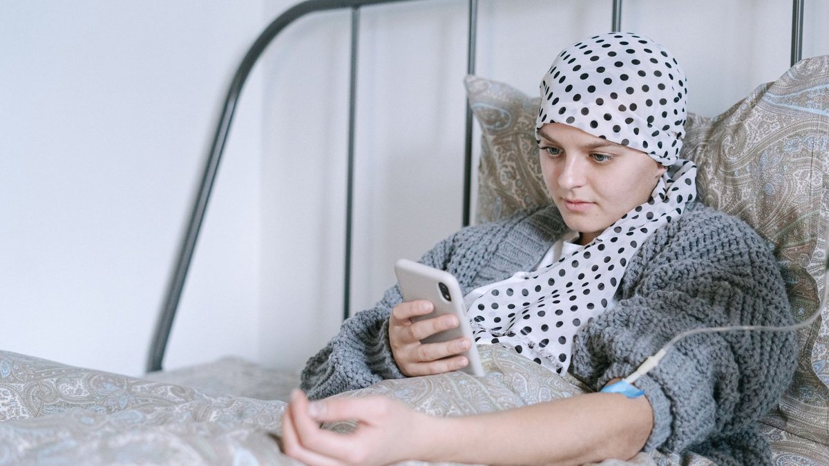 Woman in bed undergoing cancer treatment, looking at her phone with hope and determination.