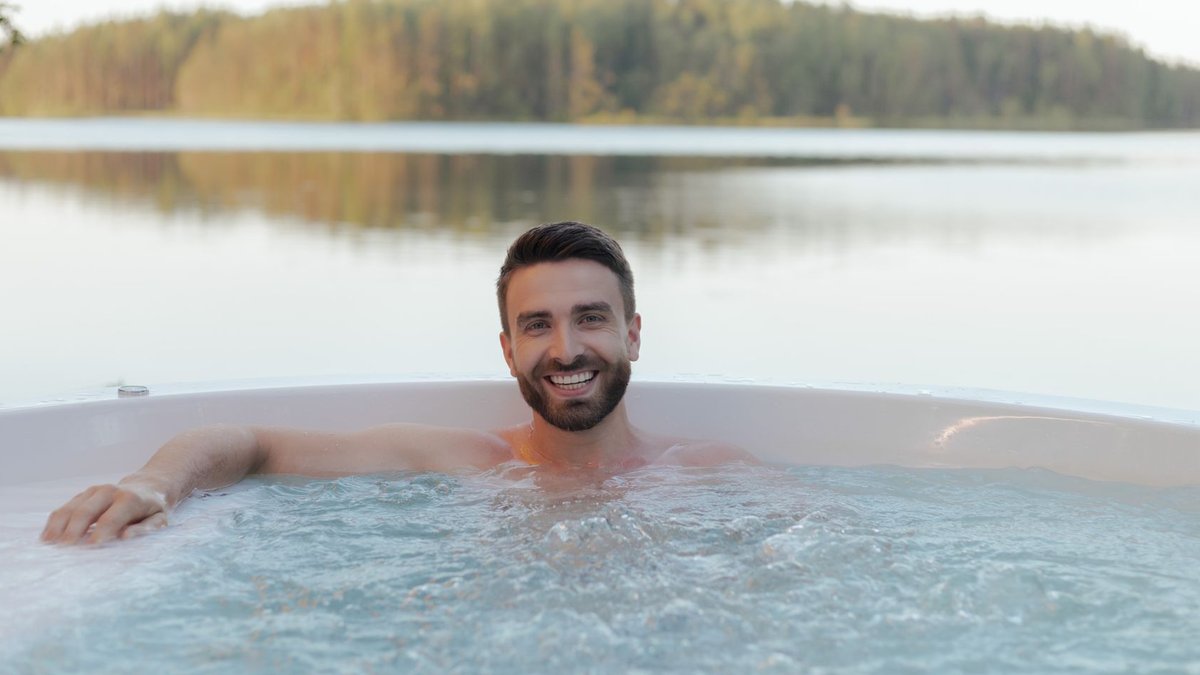 Shirtless man smiling in an outdoor hot tub by a serene lake, enjoying relaxation and wellness.