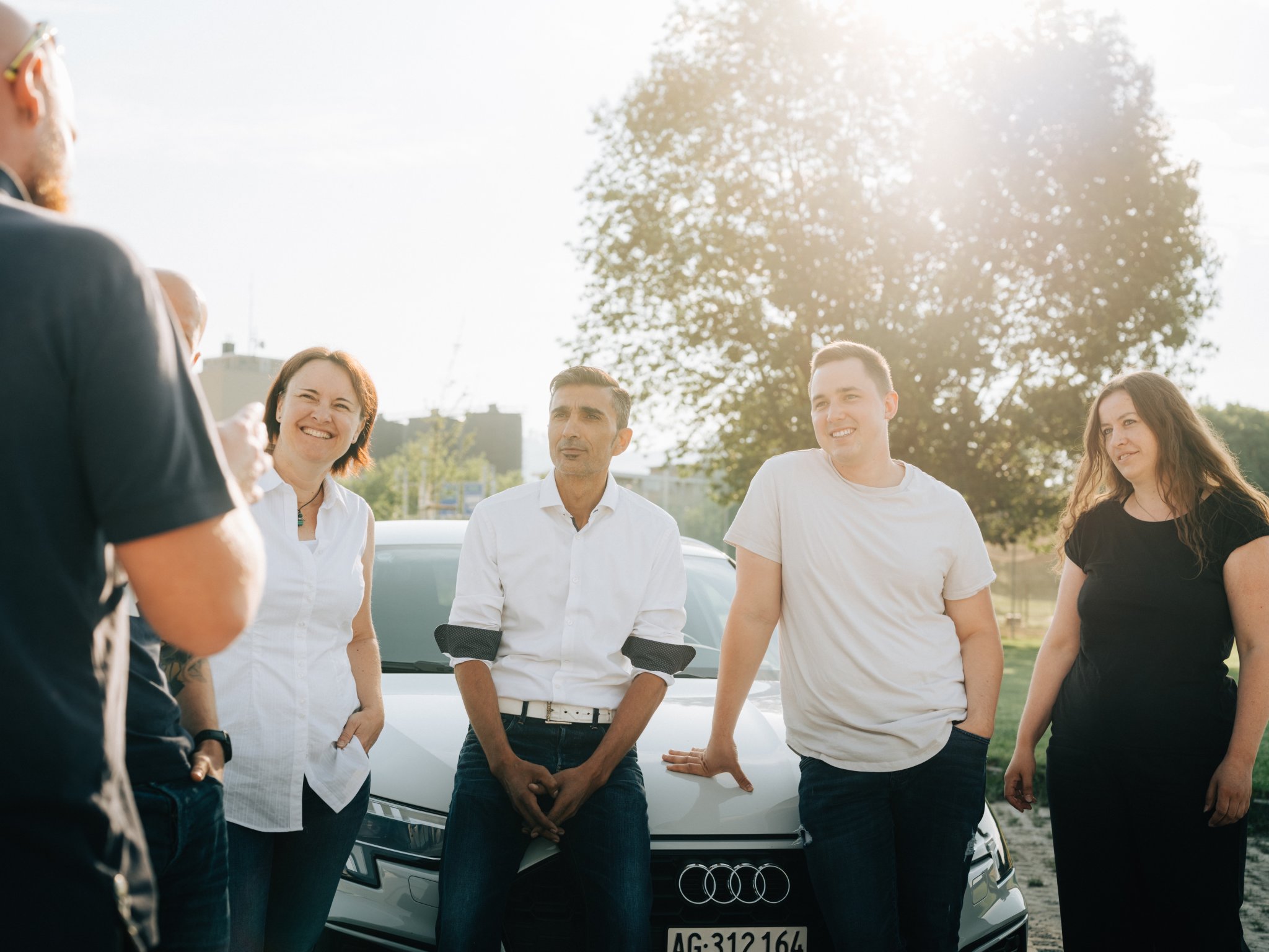 Fahrlehrer:innen von BLINK Frauenfeld lachen gemeinsam vor einem weissen Fahrschulauto der Marke Audi.