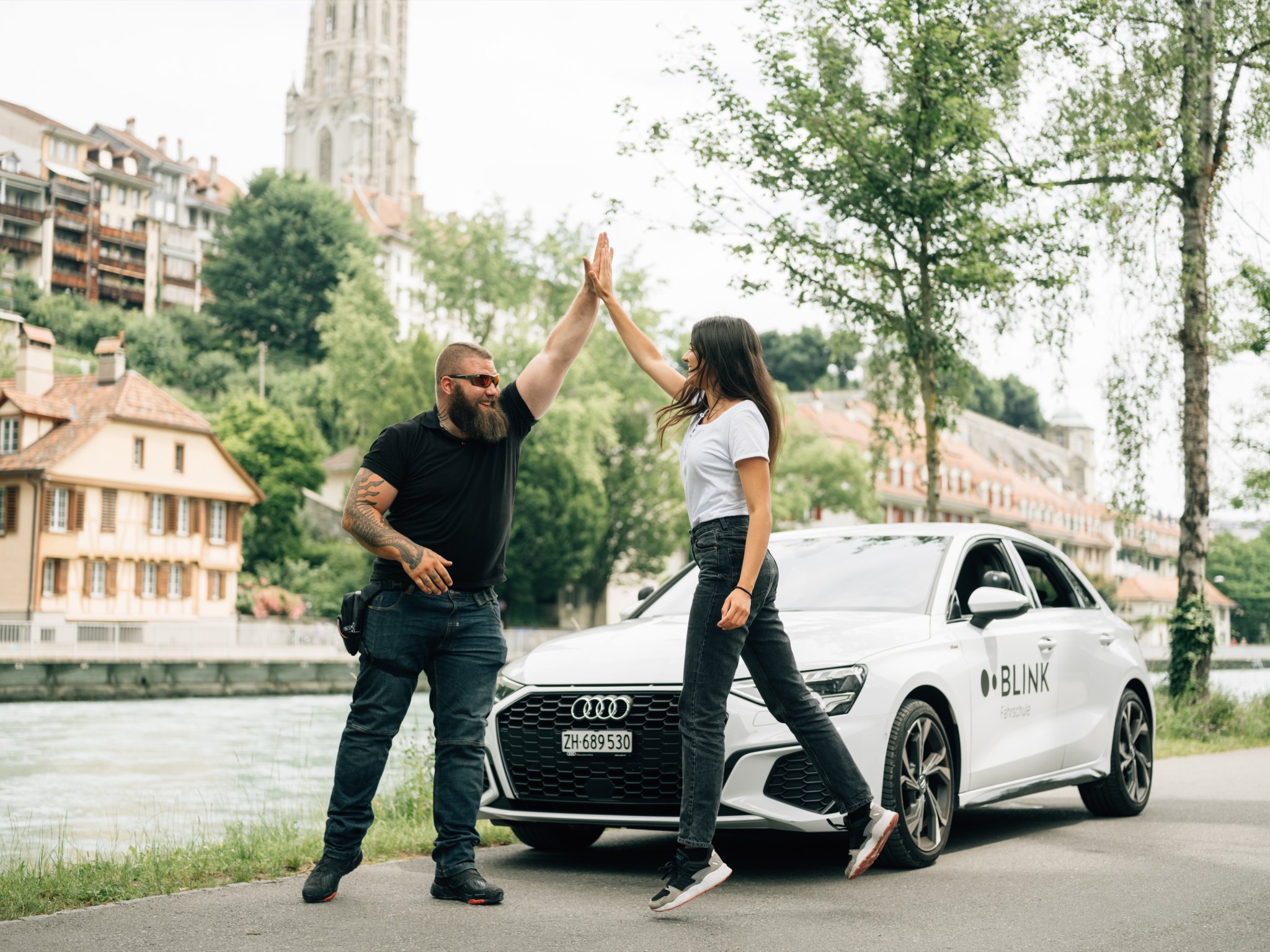 Eine Fahrschülerin und ein Fahrlehrer von BLINK stehen vor einem weissen Fahrschulauto und geben sich ein High-Five.