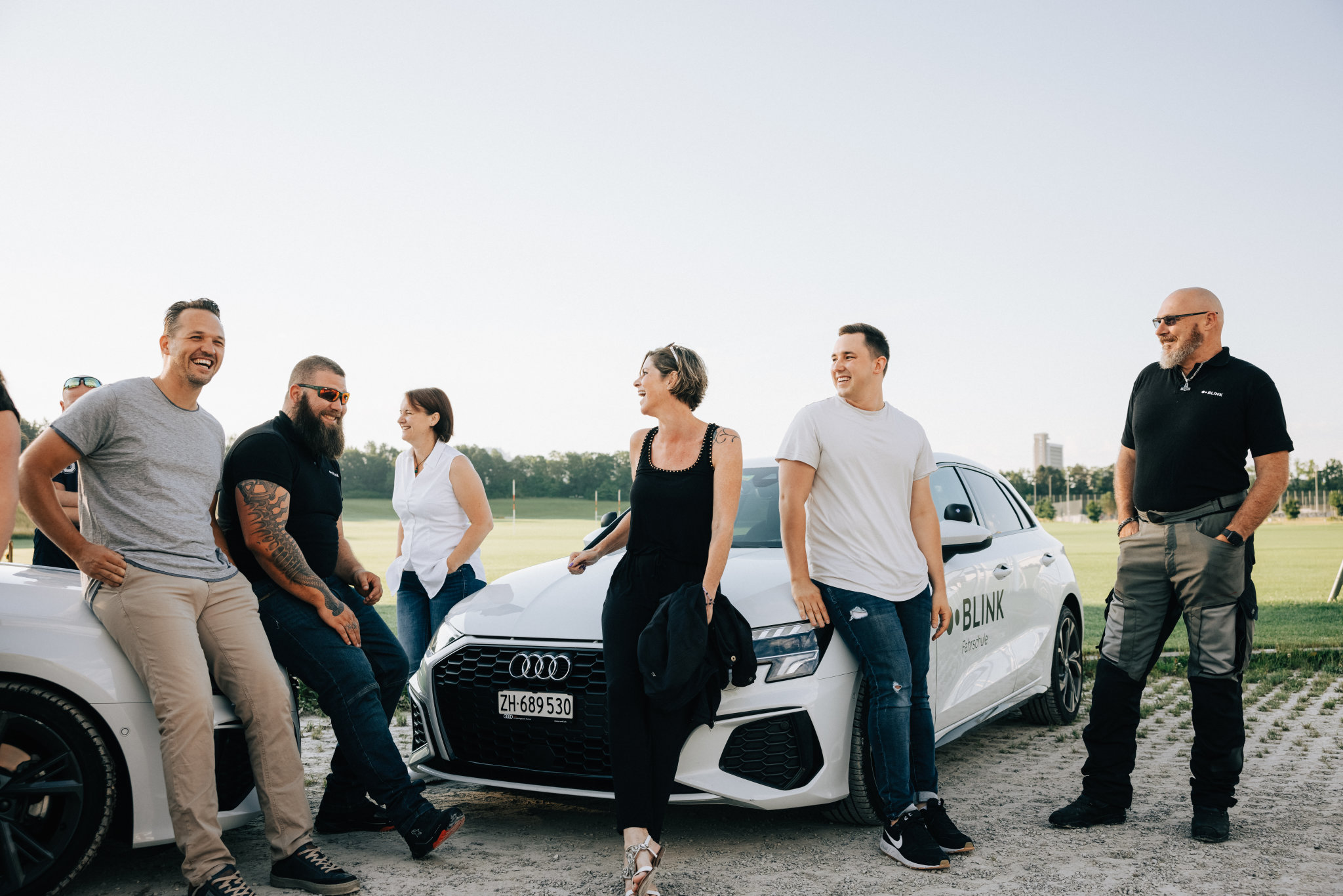 Acht Fahrlehrer:innen der BLINK Fahrschule stehen vor zwei Fahrschulautos und lachen