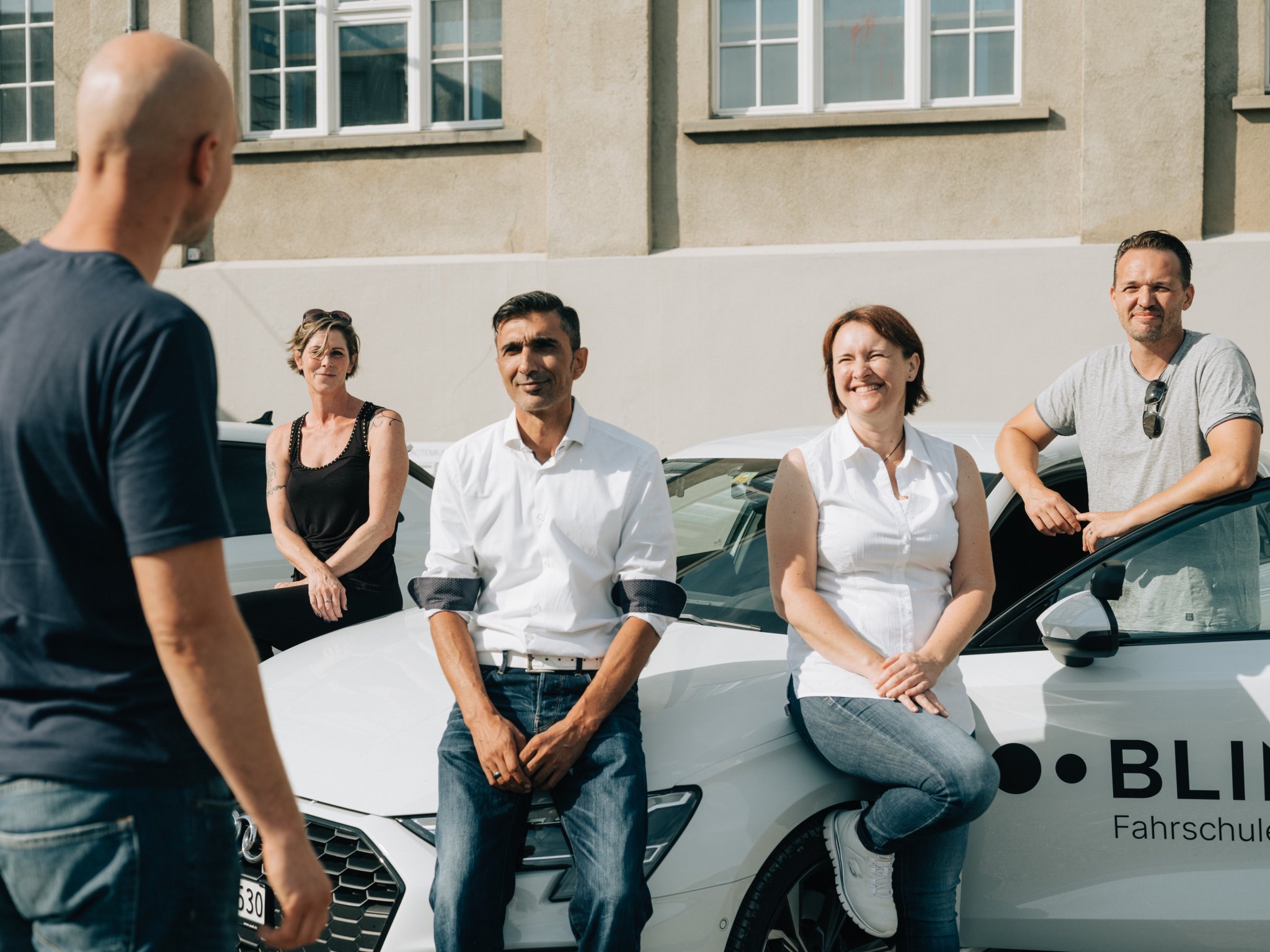 Fünf Fahrlehrer:innen der BLINK Fahrschule stehen vor einem Fahrschulauto und blinzeln nett in das Sonnenlicht.