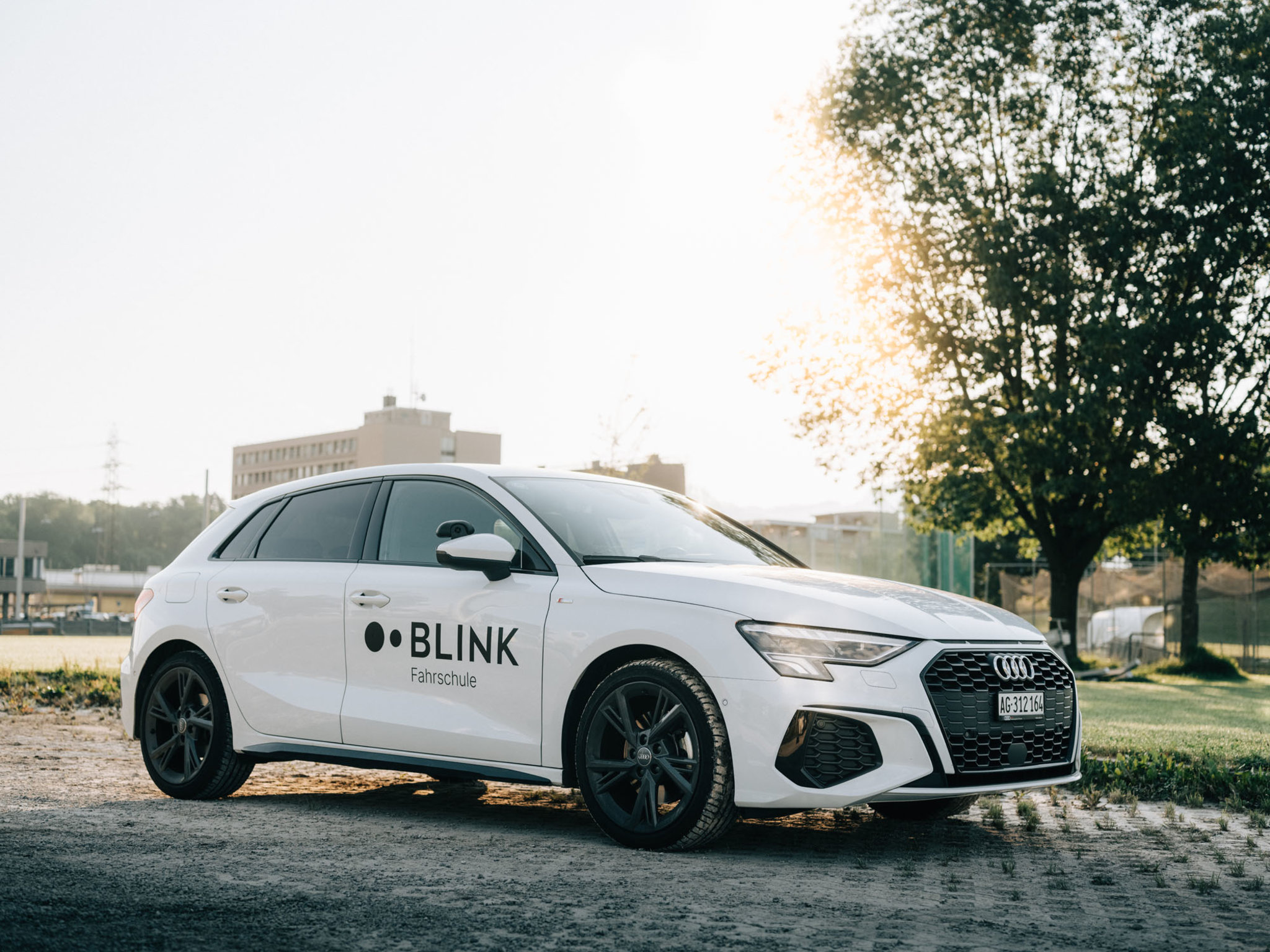 Ein weisses BLINK Fahrschulauto der Marke Audi steht auf einem grossen Parkplatz im Sonnenlicht.