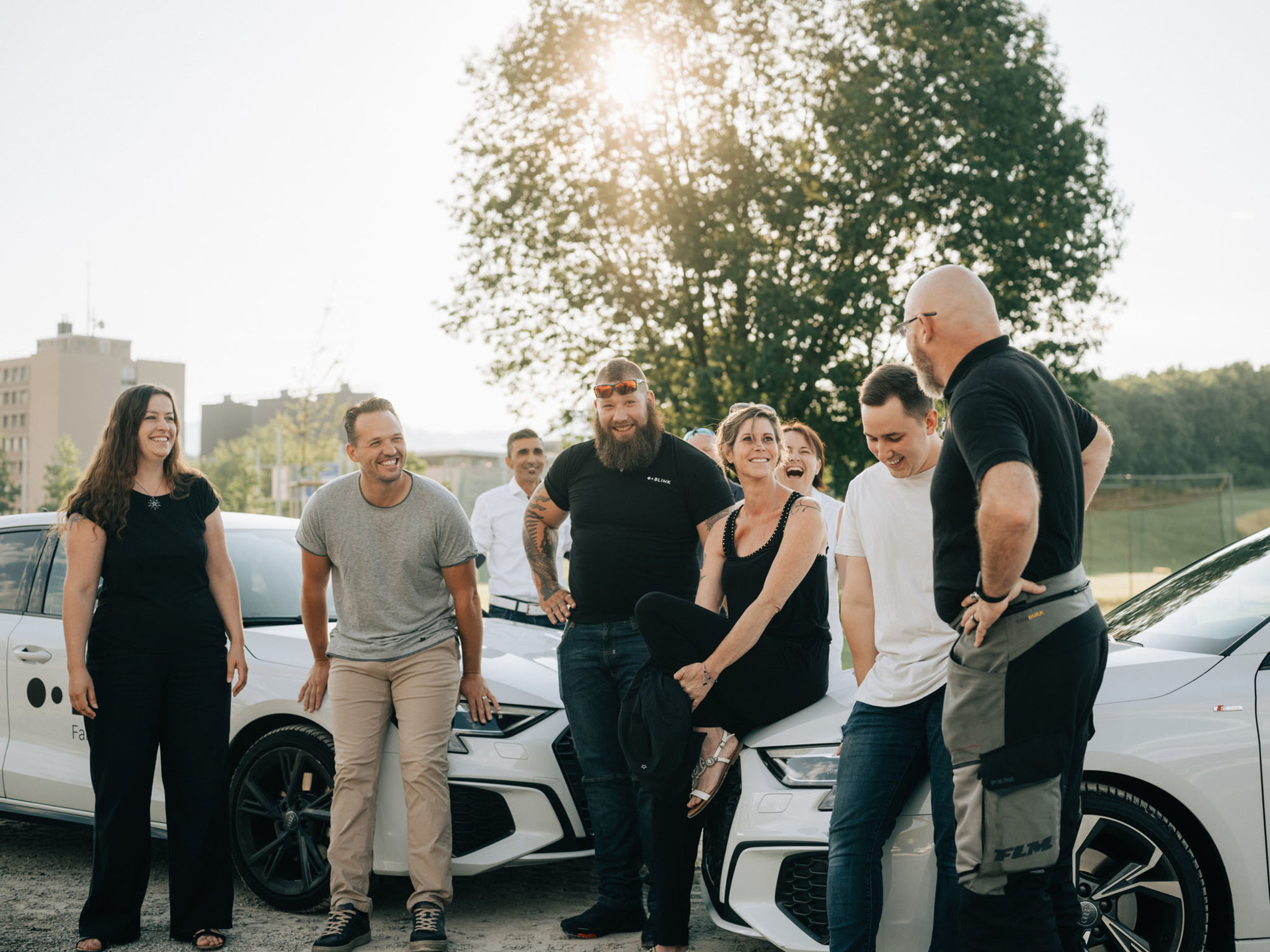 Fahrlehrer:innen aus dem BLINK Team, die sich vor zwei weissen Fahrschulautos versammelt haben und gemeinsam lachen.