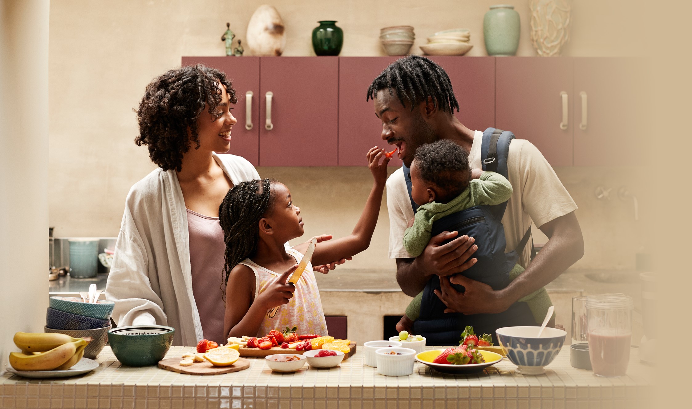 A family (mother, father, daughter and an infant in a baby-wearing sling) are in a comfortable, well stocked kitchen. The daughter offers a just-cut piece of strawberry to her father, who is hugging the infant in the baby sling he wears.