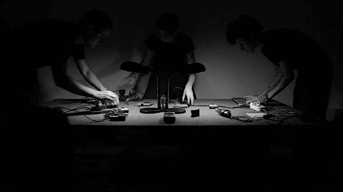 A black and white photo showing (from L-R) Hebert Baioco, Vanessa De Michelis, and Jiulian Gonçalves in a performance of The Tension in 2015 at Galeria Transitória. All three are wearing black trousers and black t-shirts, and are standing around a table in a dark room, lit by a pair of desk lamps in the center of the table. All three are operating sound mixing and looping equipment as a part of their performance.