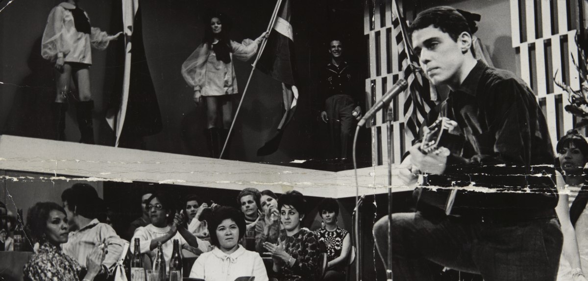 A black and white photograph of Chico Buarque performing on TV Rio from 1967. Chico Buarque is standing on the right side of the image, playing an guitar in front of a stand microphone. An audience of seated cafe goers can be seen on the left. There are two women visible on an elevated platform above and to the rear of the seated audience, they are wearing go-go boots and 1960s style mod coats.