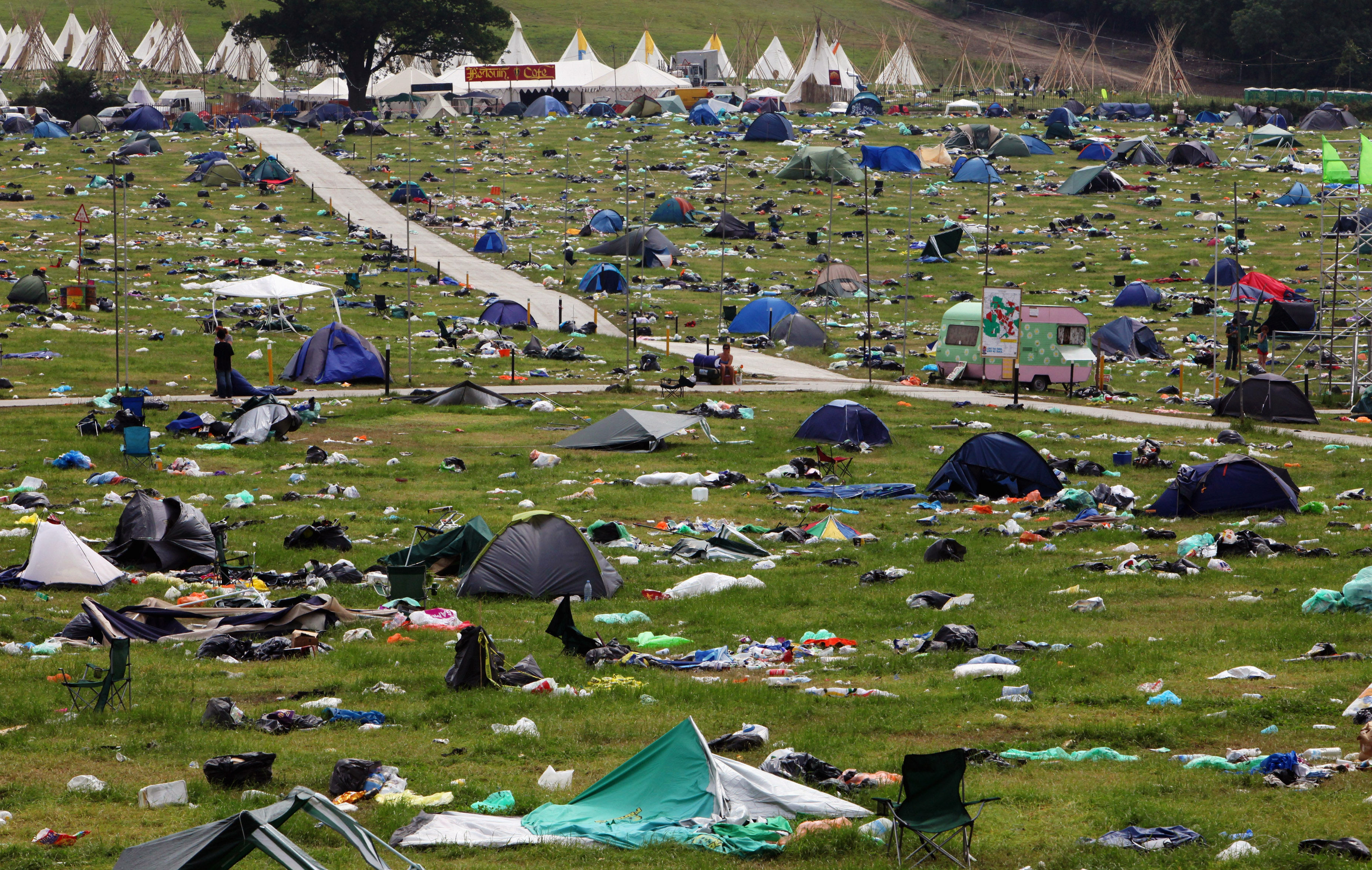 A photograph of a field at the Glastonbury farm, taken after the end of the 2009 Glastonbury Music festival. The wide, grassy field is strewn with abandoned and collapsed tents, damaged camp chairs, and countless heaps of garbage, carrier bags, empty cartons and other post-festival debris. At the edge of the field, a large, multi-pointed marquee tent is visible. A few, isolated people can barely be seen in different parts of the field.