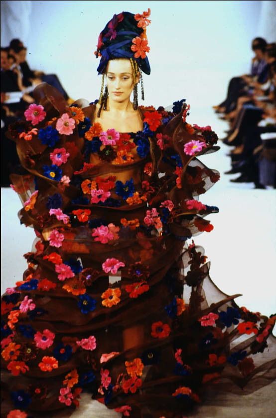 Runway model wearing voluminous sheer floral gown with colorful flower embellishments