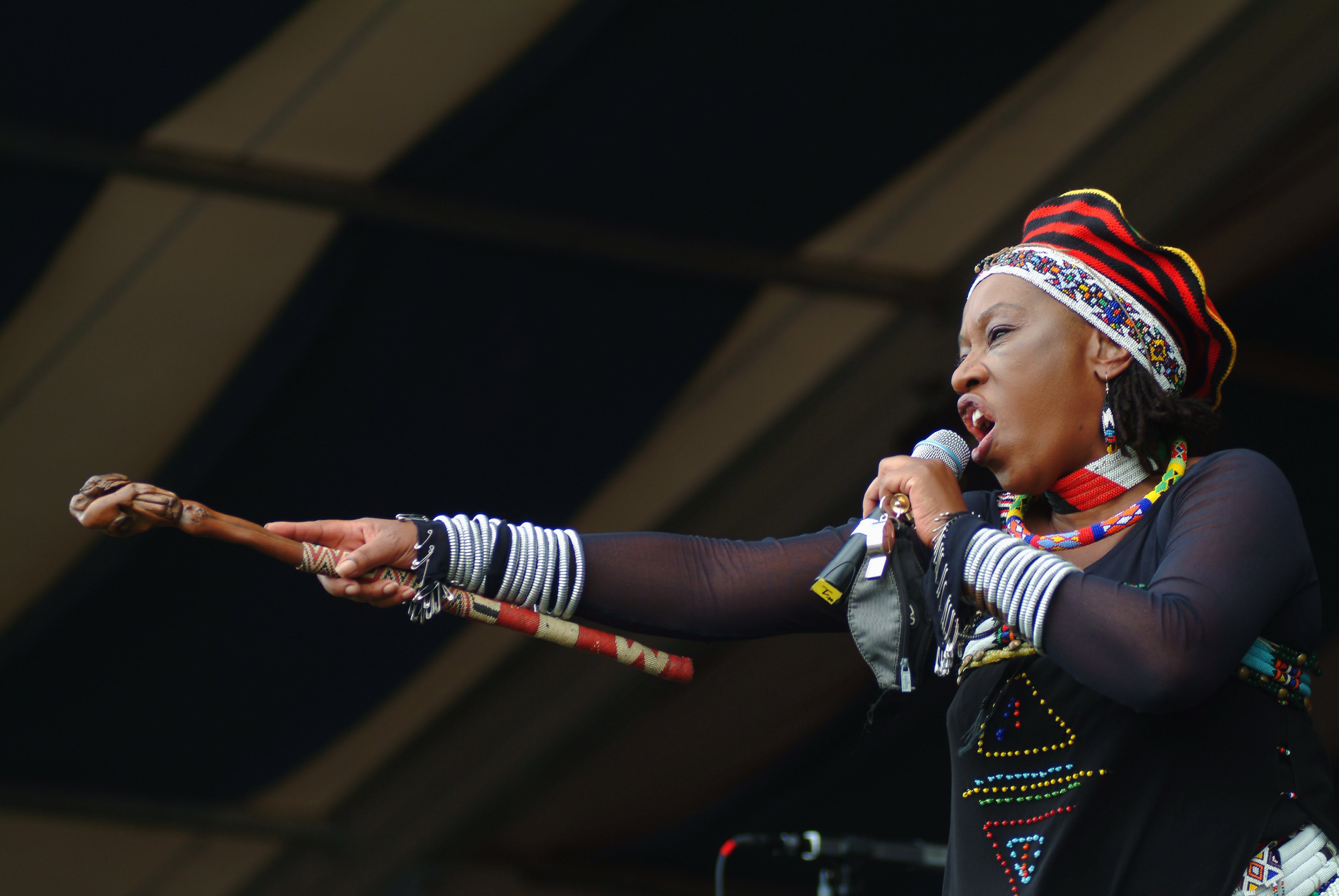 A photo of Maskanda artist Busi Mhlongo holding out a stick and singing into a microphone.