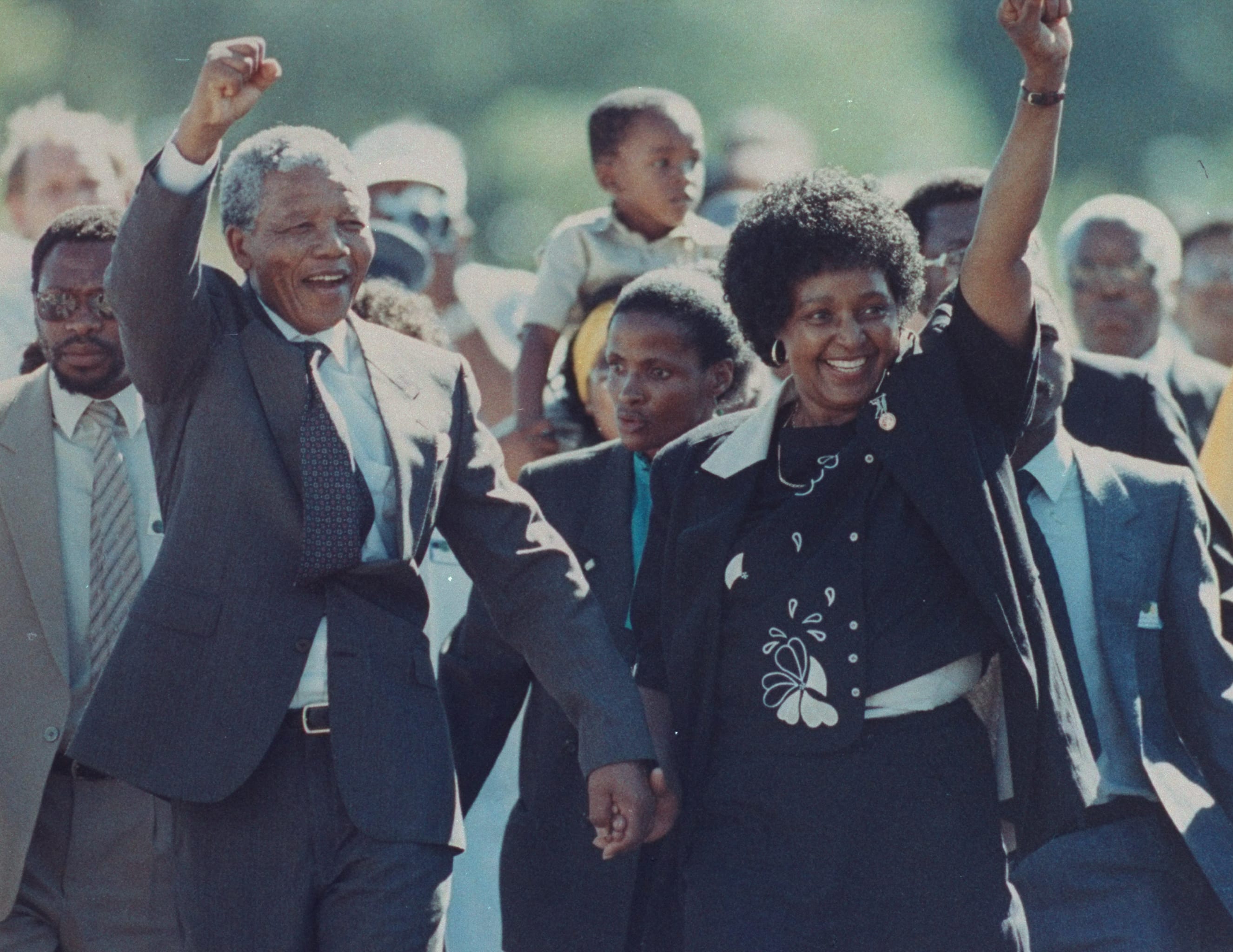 A photo of Nelson Mandela and his wife Winnie raising their fists upon his release from Victor Verster prison after 27 years. They are outside and have people behind them joining hands.