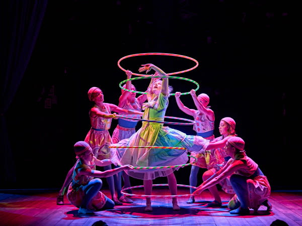 A scene from the stage production Ballet Shoes. A young woman in a wide skirted green dress reaches and looks upwards. She stands within several colourful striped hula hoops  held by other members of the cast. Credit Line: Ballet Shoes Photo by Manuel Harlan