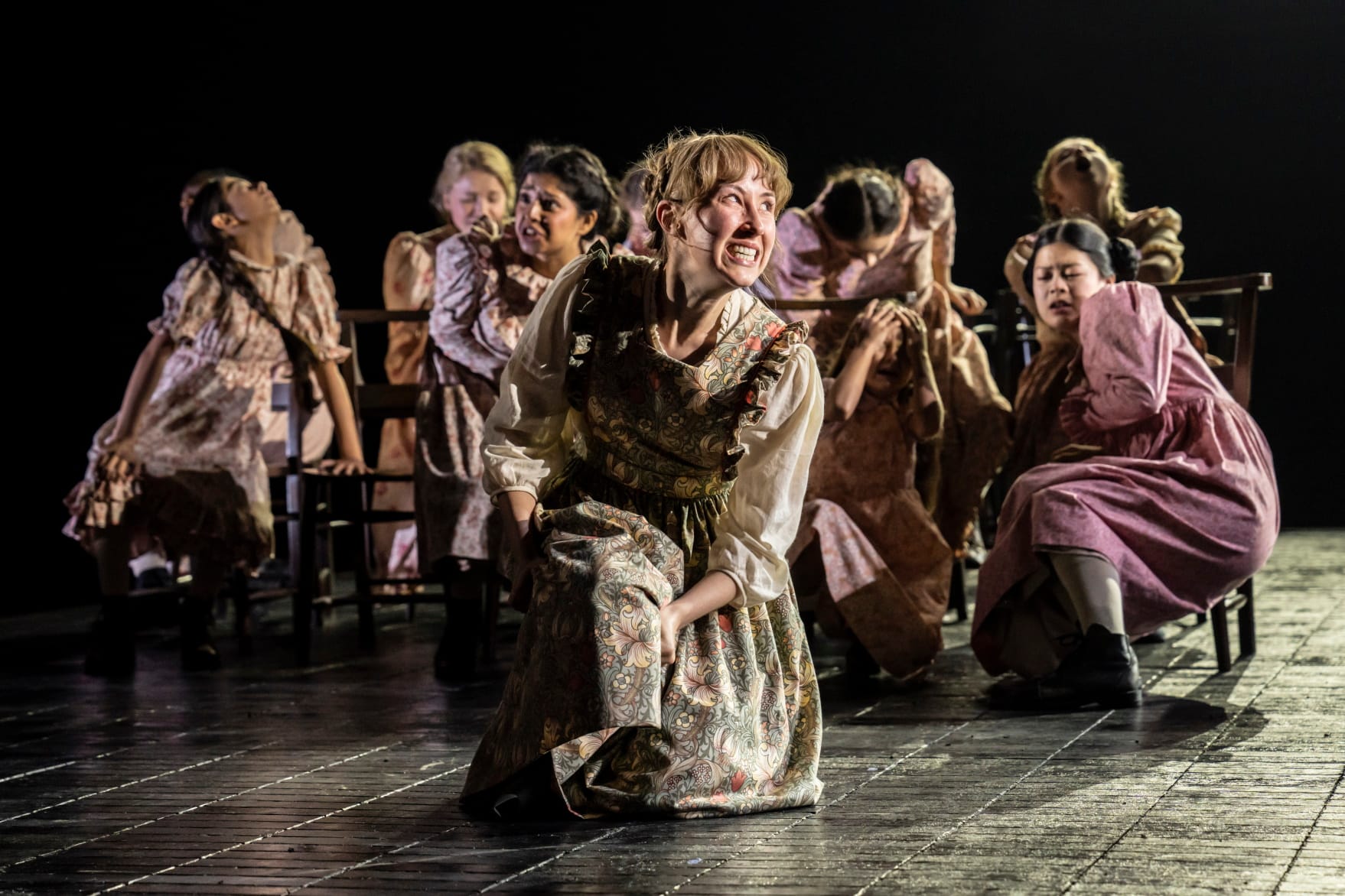 A group of young women in old-fashioned pinafores and dresses crouch on the stage with expressions of intense pain or discomfort, in a production of The Crucible by Arthur Miller, directed by Lyndsey Turner. Credit line: The Crucible photo by Johan Persson