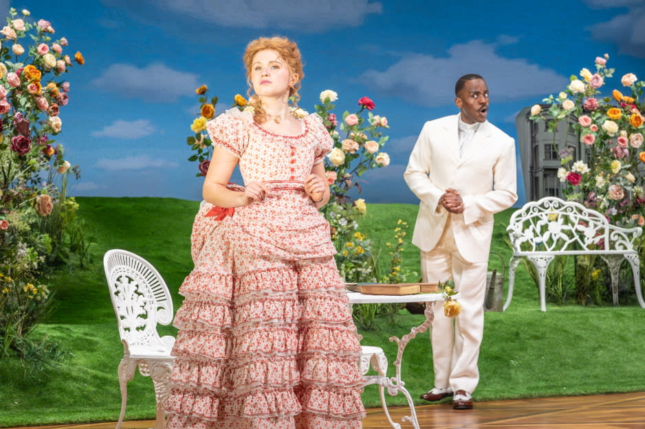 A woman in a pink floral Victorian dress with ruffled layers (portraying Gwendolen) stands in the foreground holding a letter, looking thoughtful. Behind her, a man in a white suit (played by Ncuti Gatwa as Algernon) stands near a white garden bench, looking toward her with a surprised expression. Credit line: The Importance of Being Earnest photo by Marc Brenner