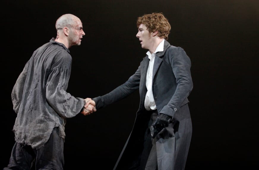 Benedict Cumberbatch and Jonny Lee Miller on stage during a production of Frankenstein. They both wear dark clothes against a black background and they stand in profile opposite each other, grasping right hands. Credit line: FRANKENSTEIN Photo by Catherine Ashmore
