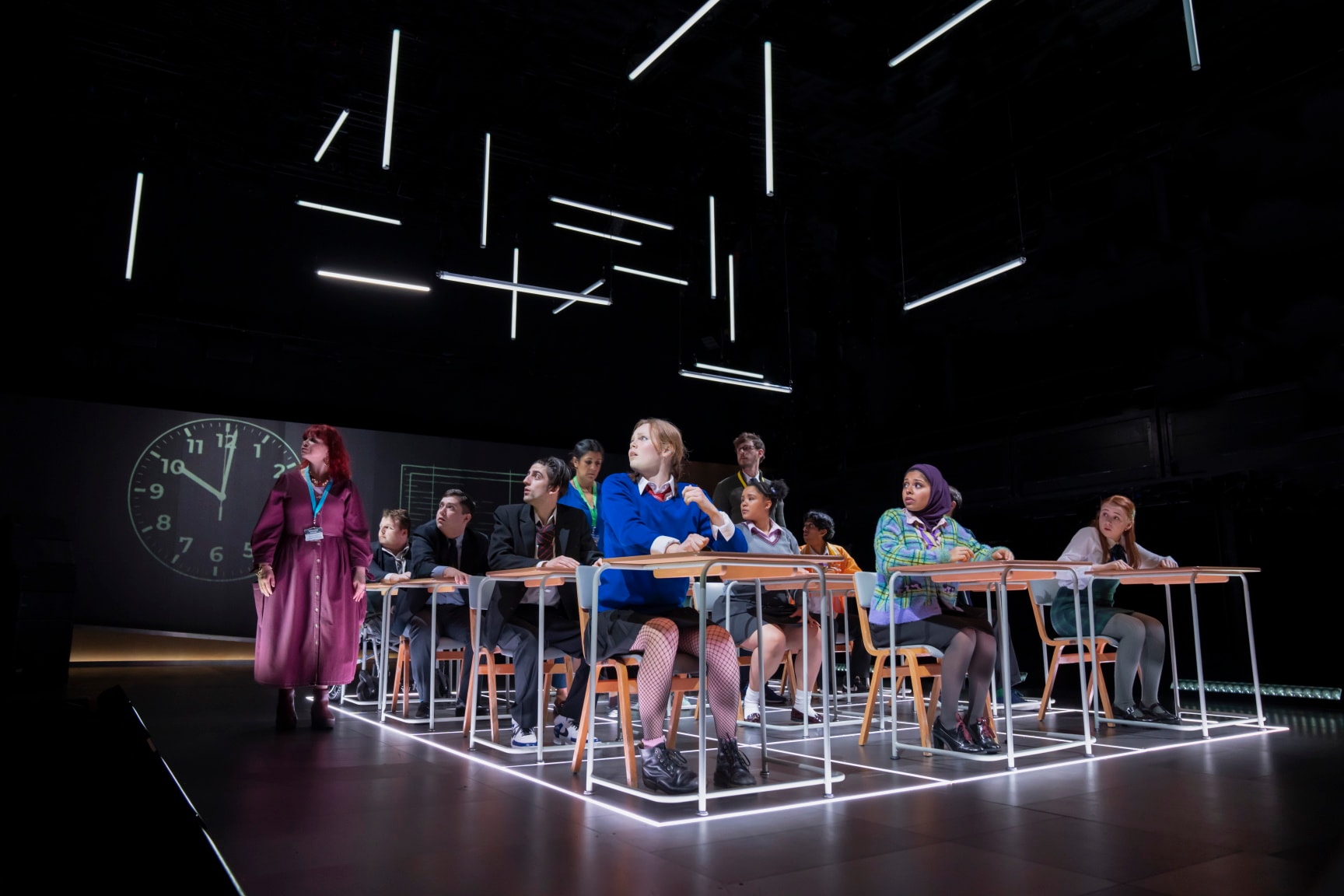 A scene from the stage production of Our Generation. A class of school children sit at old fashioned wooden and metal school desks looking to the left of the stage with concerned expressions. Three adults wearing lanyards stand towards the back of the class, also looking to the left, with a large clock visible in the background. Credit line: Our Generation photo by Johan Persson