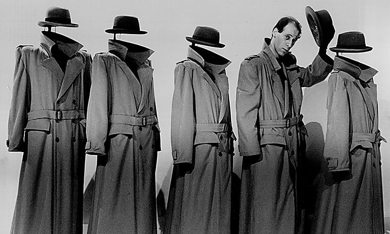 A man in a trench coat doffs his hat whilst in a row of mannequins with trench coats and hats