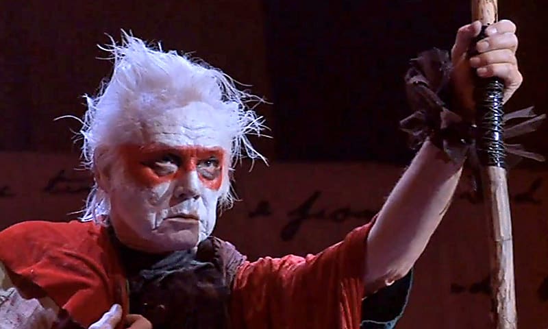 An actor dressed in costume with white and red face paint holding a stick for the Shakespeare Video Collection