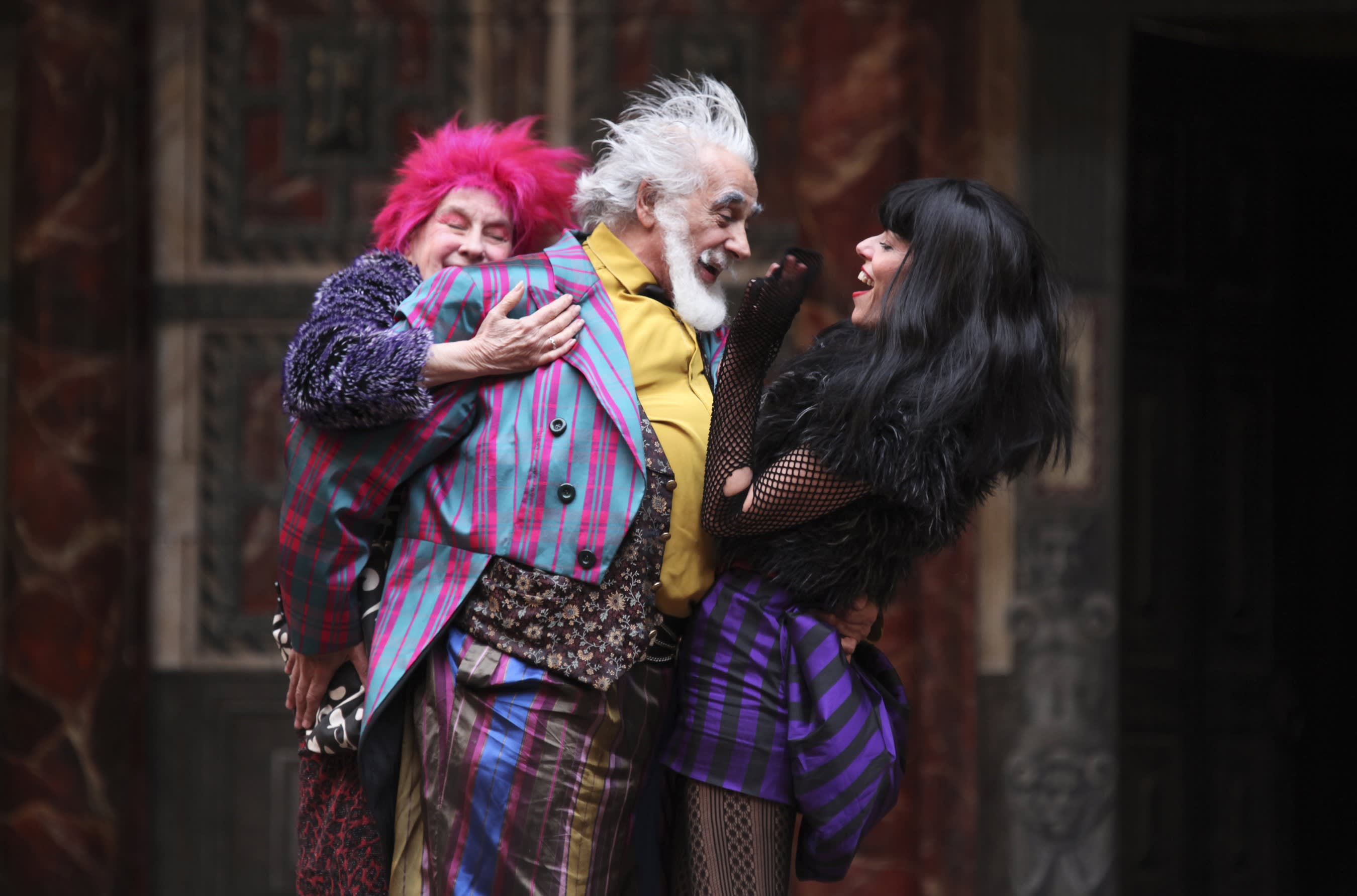 A white-haired man with a beard wearin a pink and blue striped suit has his arm around a dark-haired woman wearing black and purple clothes, the man is also being hugged by another woman