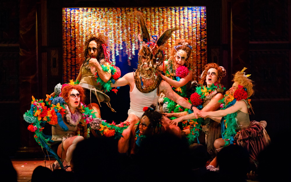 An actor with a large brown puppet donkey mask plays Bottom in A Midsummer Night's Dream and is surrounded by actors with bright flower wreaths and pom poms